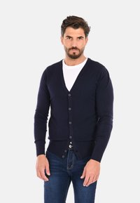 Cardigan a maglia blu navy con scollo a V, maniche lunghe, chiusura frontale a sei bottoni e orlo a coste; indossato sopra una camicia bianca.