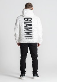 Bijela puffer jakna s quiltiranim uzorkom, s crnim logotipom na leđima. Upari s crnim hlačama i crnim tenisicama.