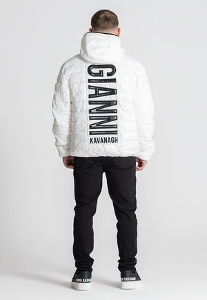 Chaqueta de plumas blanca con textura acolchada, que presenta un texto de logo negro en la parte trasera. Combinada con pantalones negros y zapatillas negras.