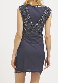 Tmavě modré šaty bez rukávů z průsvitné látky, zdobené stříbrnými flitrovými akcenty ve tvaru křížového vzoru na zádech.