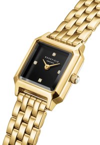Reloj rectangular en tono dorado con esfera negra, que cuenta con cuatro indicadores de horas similares a diamantes, manecillas doradas y una pulsera de eslabones.