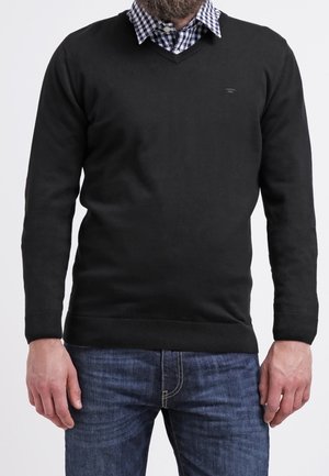 Homme portant un pull noir à col en V par-dessus une chemise à carreaux bleue et blanche assortie d'un jean bleu, debout devant un fond uni.