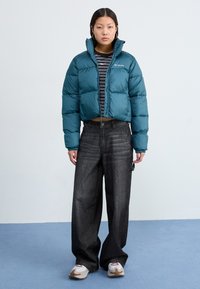 Teal pufferjakke med quiltet design, høy krage og logo. Brukt over en stripete skjorte og kombinert med vide grå jeans og sneakers.