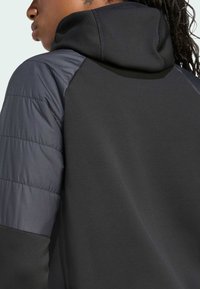 Zatvoren kadar ramena i vrata osobe, s crnom kapuljačom i sivim kviltanim rukavom, naglašavajući teksture i boje tkiva.