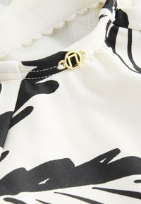 Gros plan sur un tissu à motif noir et blanc avec un petit logo rond en métal doré près d'une couture cousue.