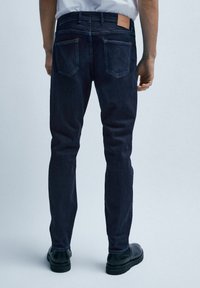 Mörka denimjeans med slim fit, utrustade med bakfickor och en brun läderetikett på midjebandet. Materialet verkar robust och strukturerat.