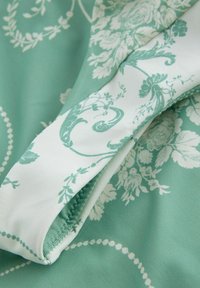 Šviesiai žalia medžiaga, puošta baltomis gėlėmis ir scrollo raštais. Lygus paviršius su kontrastinga balta apdaila kraštuose.