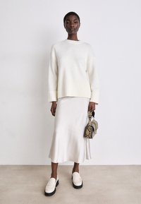White knit sweater with ribbed hem, paired with a satin cream skirt. White loafers with black soles and a small patterned handbag.