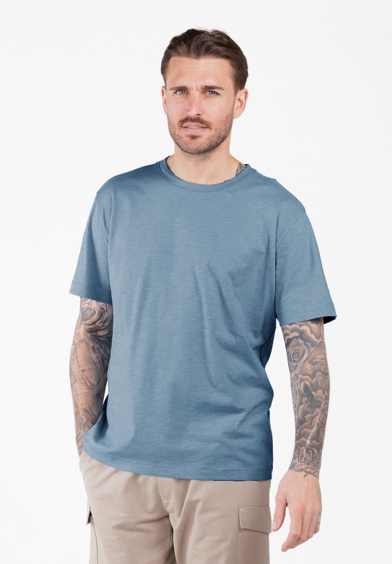 Mann mit kurzen braunen Haaren und Tätowierungen an beiden Armen, trägt ein blaues kurzärmeliges T-Shirt und beige Hosen mit den Händen in den Taschen.