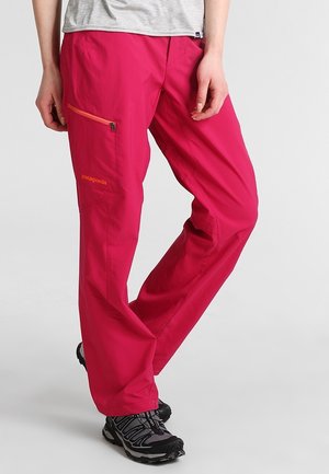 Persoon draagt felroze outdoorbroek met oranje ritszak en zwarte wandelschoenen, staand tegen een effen achtergrond.