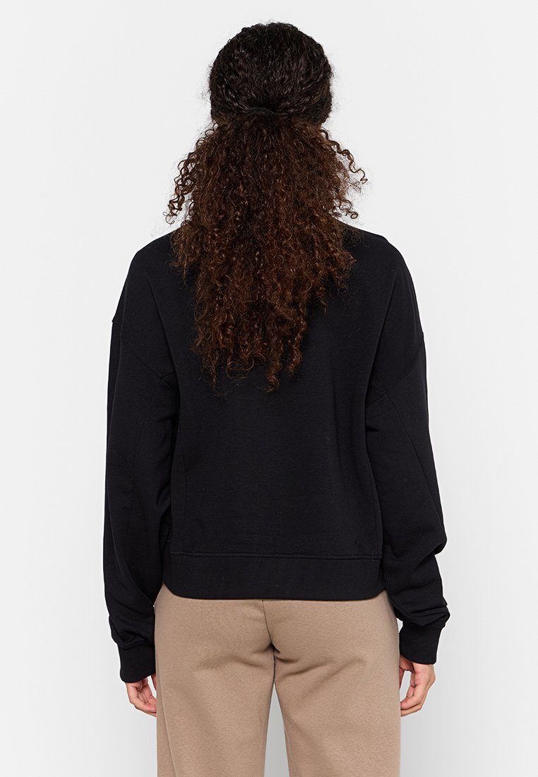 Sudadera negra con un corte relajado, puños y dobladillo acanalados. Textura suave, mangas anchas. Llevada con pantalones beige, vista desde la parte posterior.