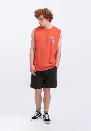 Rode mouwloze T-shirt met een klein grafisch detail, gecombineerd met zwarte korte broek en zwarte sneakers. Een casual en sportieve outfit.