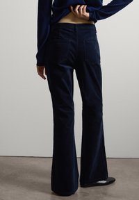 Persona vistiendo pantalones de pana azul oscuro y zapatos negros, de pie con una mano detrás de la espalda frente a un fondo liso.