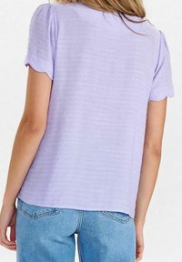 Donna che indossa una blusa a maniche corte di un leggero viola con bordi ondulati e jeans a vita alta di un blu chiaro, vista da dietro.