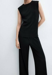 Žena v černém bezrukávovém žebrovaném topu a odpovídajících širokých žebrovaných kalhotách stojí před jednoduchou světle barevnou stěnou.