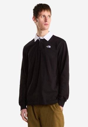 Ung mand med kort hår iført en sort langærmet poloshirt med hvid krave og brune bukser, som kigger til siden.