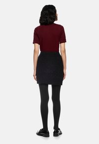 Burgunderfarbenes Kurzarmoberteil, kombiniert mit einem strukturierten schwarzen Minirock. Das Model trägt schwarze Strumpfhosen und glänzende schwarze Schuhe und steht mit dem Rücken zur Kamera.