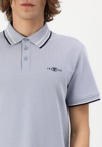 Grå polo t-shirt lavet af tekstureret stof, med en tre-knaps knaplukning, stribet krave i marineblå og lys grå, samt en lille logo detalje.