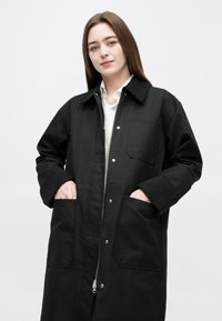 Palton negru supradimensionat cu fermoar frontal, accente cu nasturi, două buzunare frontale și guler, realizat dintr-o țesătură fină cu manșete tricotate.