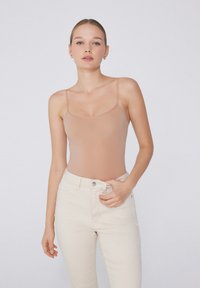 Tělový bodystocking s tenkými ramínky a bezešvým designem; spárován s vysokým pasem, světle béžovými džíny. Jednoduchá, hladká textura.