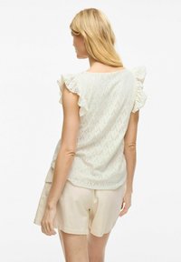 Hvid bluse med broderet hulmønster og voluminøse ærmer, prydet med et diamantmønster, der suppleres af beige shorts. Den strukturerede stof giver visuelt interesse.