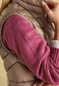 Puffige beigefarbene Weste über einem rosa gestrickten Pullover mit strukturiertem Stoff. Der linke Arm zeigt Rippendetails und ein weiches Strickmuster.