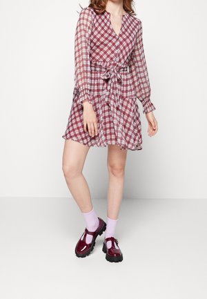 Sukienka kopertowa w kratę w odcieniach czerwieni z długimi rękawami i wiązaniem w talii. Modelka ma na sobie błyszczące bordowe buty i jasnoróżowe skarpetki.