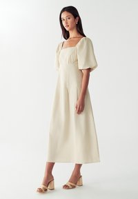 Svijetlosmeđi midi haljina s kvadratnim izrezom, napuhanim rukavima i prilagođenim tijelom. Glatka tkanina s blagom teksturom. Uparena s tamnosmeškim sandalama na blok petu.