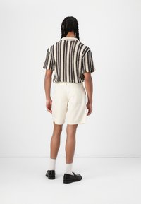 Chemise à manches courtes rayée en noir et blanc, associée à un short crème. Modèle portant des chaussures noires et des chaussettes blanches, vu de derrière.