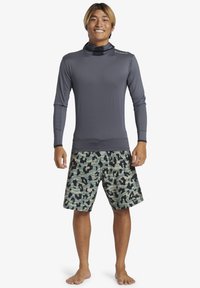 Långärmad grå rash guard med hög krage och tumhål, tillsammans med gröna camouflage-simshorts med svarta mönster och mesh-detaljer.