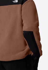 Fleece-tröja i rostbrun med en svart överdel. Har en mjuk textur, elastiska muddar och justerbar nederkant med snodd.