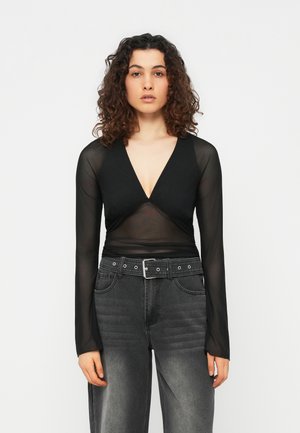 Γυναίκα με κατσαρά μαλλιά, που φοράει μαύρο διάφανο τοπ με μακριά μανίκια και βαθύ V-λαιμόκοψη, καθώς και ξεθωριασμένο μαύρο τζιν ψηλόμεσο με ζώνη.