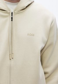 Beżowa bluza z kapturem zapinana na zamek z przodu, charakteryzująca się miękką teksturą i wytłoczonym logo. Projekt zawiera sznurki ściągające oraz kieszenie kangurki.