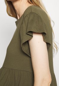 Oliwkowa bluzka z krótkimi, falbanastymi rękawami, wykonana z miękkiej bawełny, z marszczonym dołem. Widok z boku pokazuje detale rękawów.
