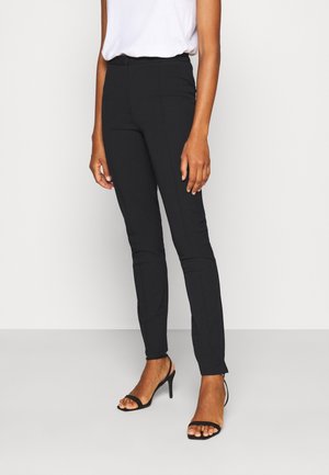 Žena v černých slim-fit kalhotách, bílém topu zasunutém do kalhot a černých otevřených vysokých podpatcích, stojící na jednobarevném pozadí.