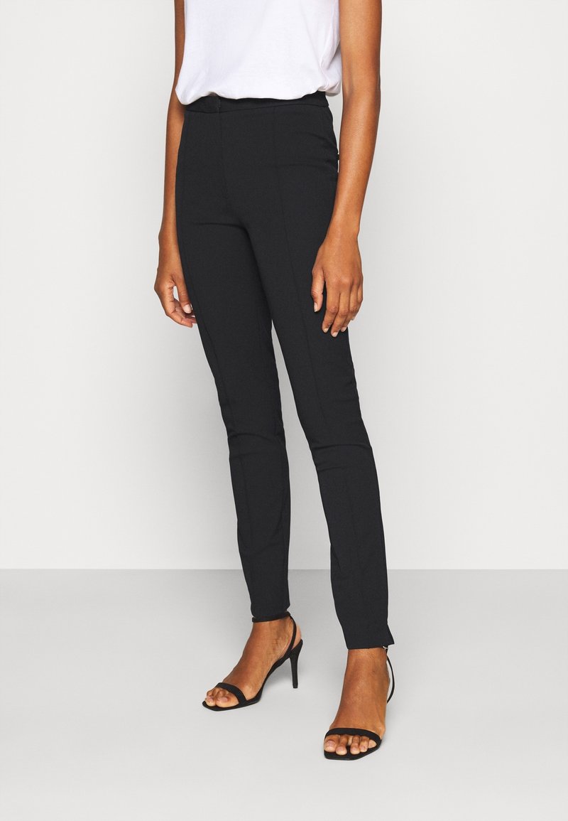Žena v černých slim-fit kalhotách, bílém topu zasunutém do kalhot a černých otevřených vysokých podpatcích, stojící na jednobarevném pozadí.