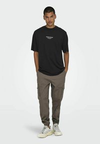 Camiseta de algodón negra con texto blanco, combinada con pantalones cargo de corte holgado en color beige oscuro, que tienen bolsillos laterales y puños elásticos, y zapatillas ligeras.