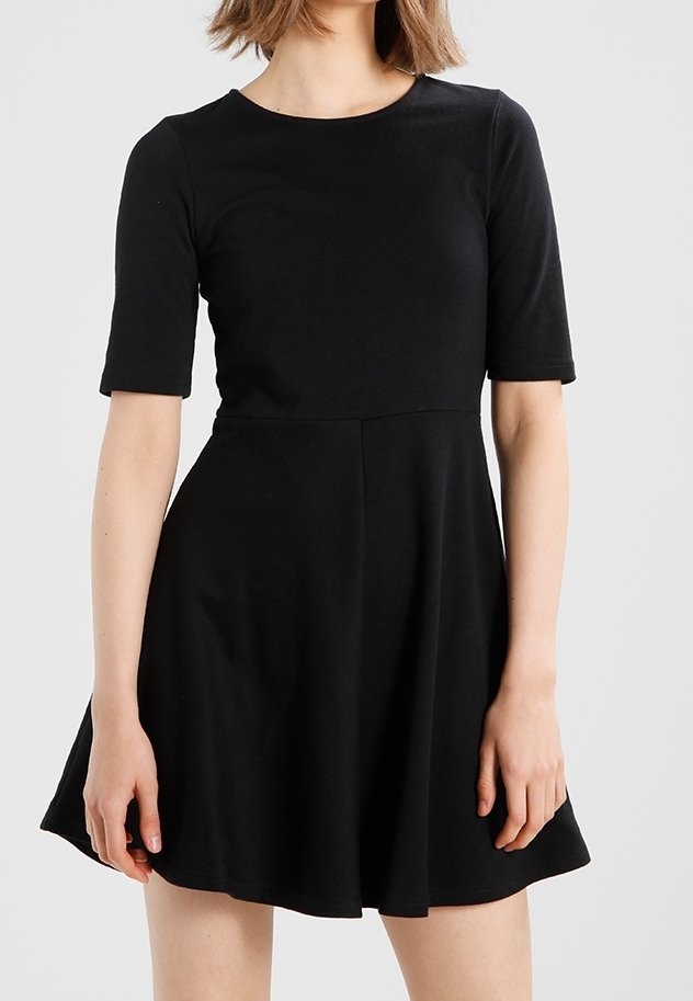 Femme portant une robe noire courte avec des manches trois-quarts et une taille cintrée, debout devant un fond gris clair uni.