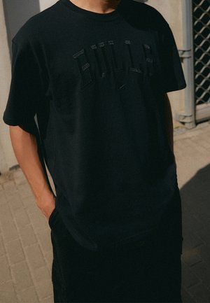 Personne portant une chemise noire à manches courtes avec un texte brodé subtil, debout à l'extérieur sur une surface pavée près d'une grille métallique.