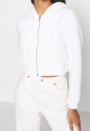 Osoba nosící bílou crop-top mikinu na zip s šňůrkami a vysokým pasem kalhoty v krémové barvě, stojící před jednoduchým pozadím.