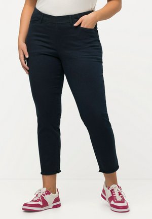 Jean skinny bleu foncé avec un ourlet effiloché, en denim, associé à des baskets blanches et roses. Comprend des poches avant et une taille ajustée.