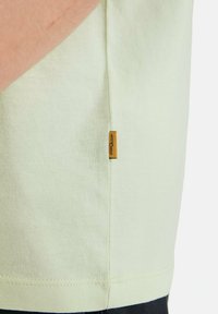 Lys grøn bomulds T-shirt med et lille gult mærke på sidesømmen, med en blød tekstur og en afslappet pasform.