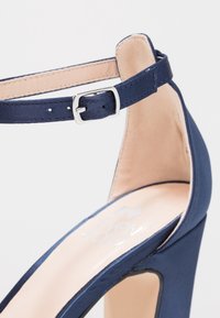 Chaussure à talon haut bleu marine avec bride à la cheville et boucle argentée, doublure intérieure beige, et finition à texture lisse.