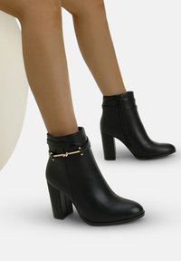 Stivaletti neri in pelle con tacco a blocco e dettaglio in oro indossati su gambe nude su uno sfondo bianco.