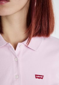 Camisa polo rosa hecha de tela texturizada, con cuello y dos botones. Parche notable del logo rojo de Levi's en el lado izquierdo.