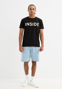 Camiseta de algodón negra con la palabra "INSIDE" impresa en blanco. Se lleva con pantalones cortos de denim azul claro y zapatillas blancas, creando un atuendo casual.