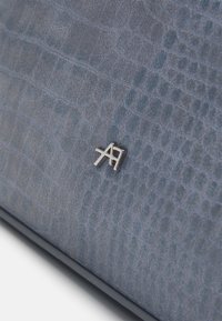 Skóra niebieska z tłoczeniem w kształcie krokodyla, o gładkiej fakturze, z metalicznym srebrnym logo. Tło stanowi jednolita, nieozdobiona powierzchnia.