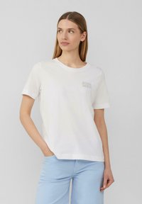 T-shirt en coton blanc à manches courtes, col rond, avec un discret texte "ÉTÉ" sur le devant. Associé à un pantalon bleu clair.