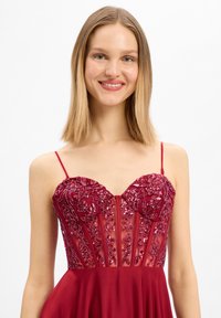 Jeune femme souriante, portant une robe rouge à fines bretelles, corsage floral pailleté et jupe fluide, devant un fond blanc uni.