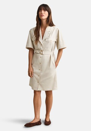 Woman wearing knee-length beige belted shirt dress with short sleeves and dark brown flats, standing against plain background.